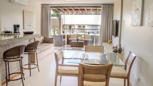 A modern living room with a glass-top dining table, two bar stools, a sofa, and a TV mounted on the wall. - Imbassaí Apartments (Mata de São João)