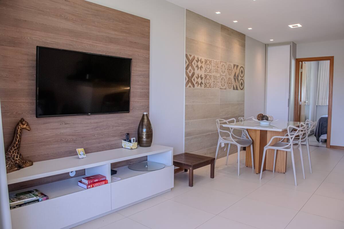 Un salon moderne avec un téléviseur à écran plat fixé à un mur en bois, un meuble de rangement blanc contenant des livres, une petite table et un coin repas avec une table en bois et des chaises blanches.