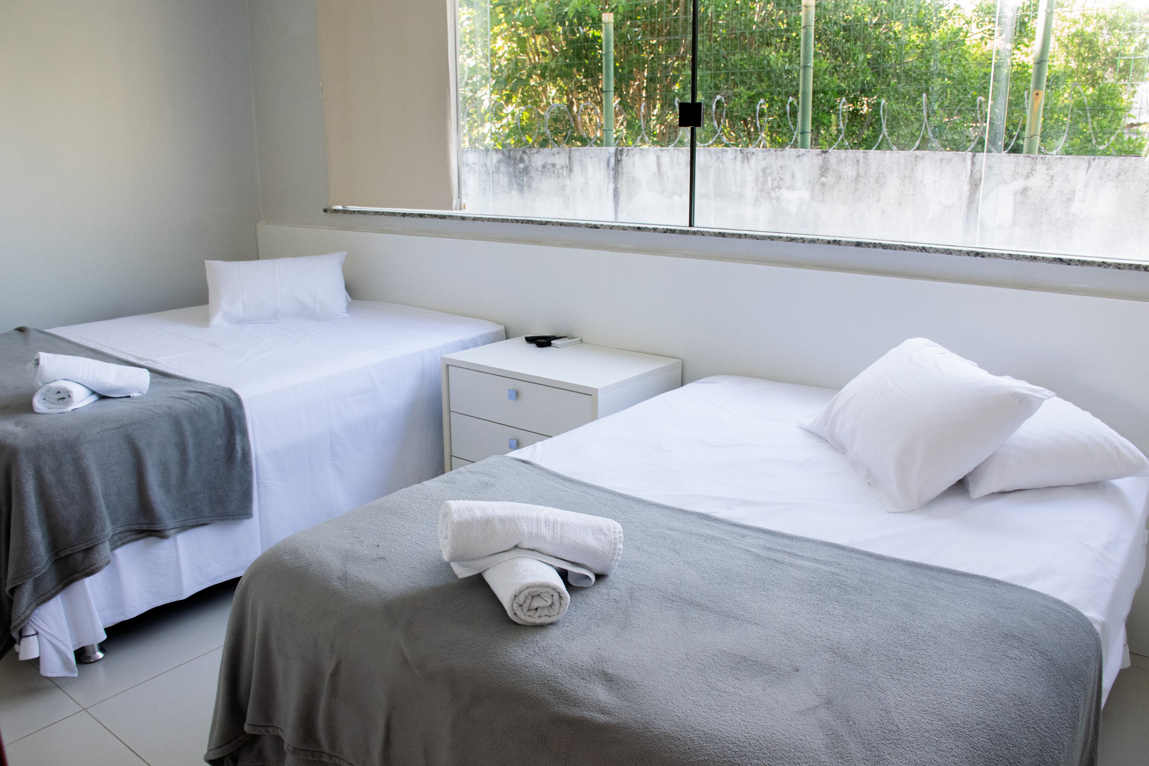 A hotel room with two beds, a small bedside table, and a window with a view of greenery.