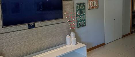 A modern living room with a large flat-screen TV mounted on a textured wall, a white entertainment unit below, and decorative wall art including a sign that says "EVERY THING IS OK".