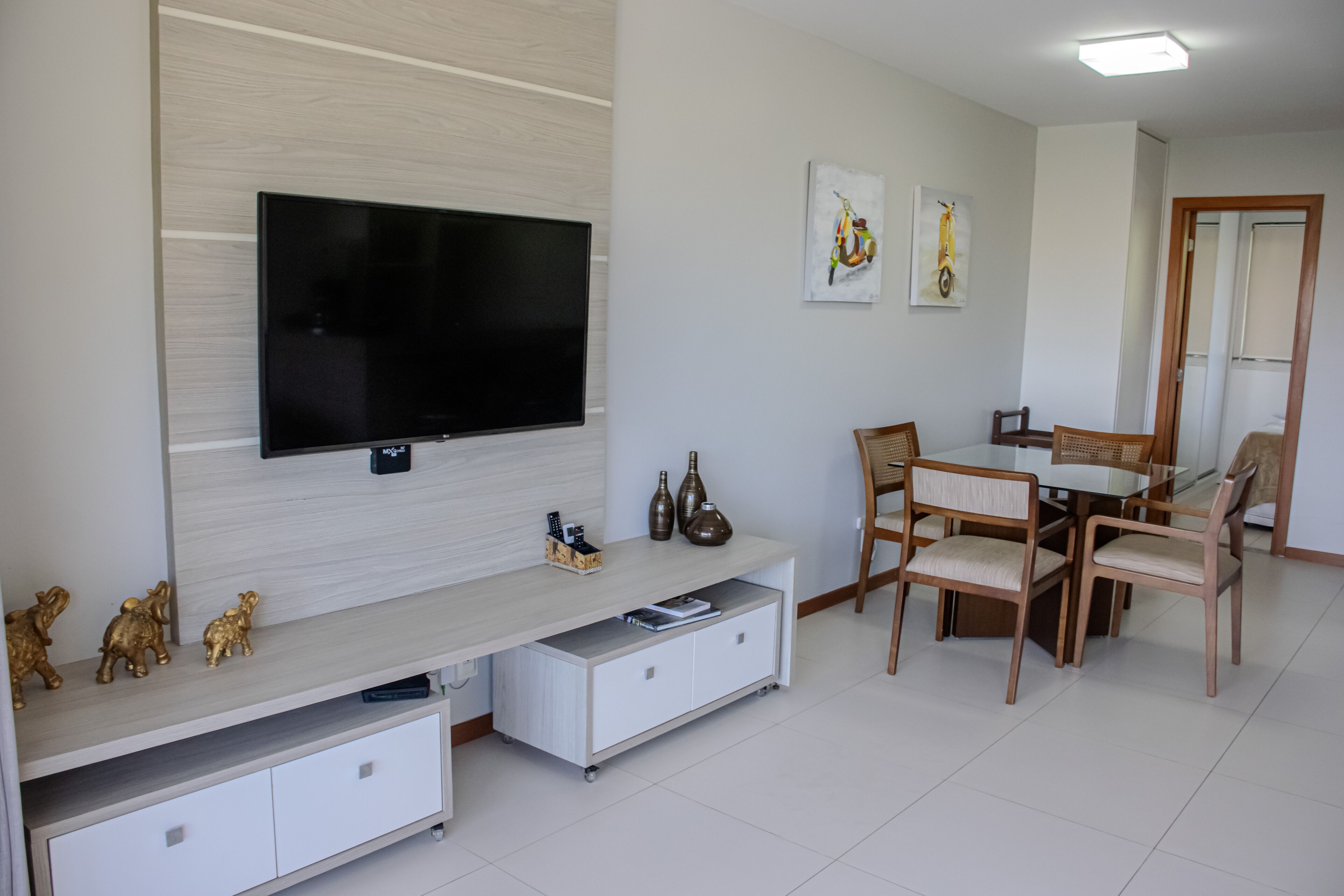 Una sala de estar moderna con un televisor de pantalla plana montado en la pared, un mueble blanco con espacio de almacenamiento, una zona de comedor con mesa y sillas, y una puerta que da a otra habitación.