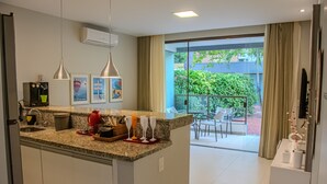 A modern kitchen with a central island, a dining area with chairs, and a view of greenery outside. - Imbassaí Apartments (Mata de São João)