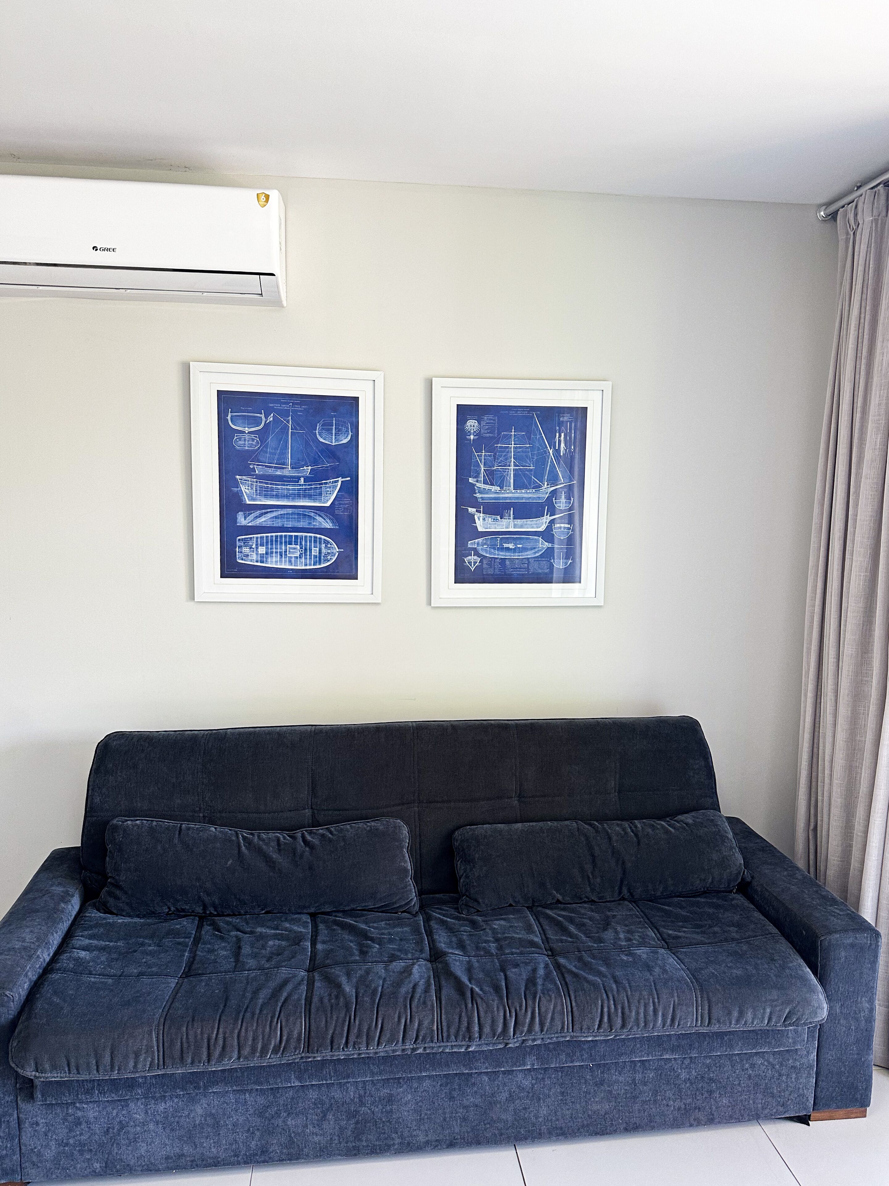 Un sofá azul oscuro con dos cojines, una unidad de aire acondicionado blanca arriba y dos dibujos arquitectónicos azules enmarcados en la pared.