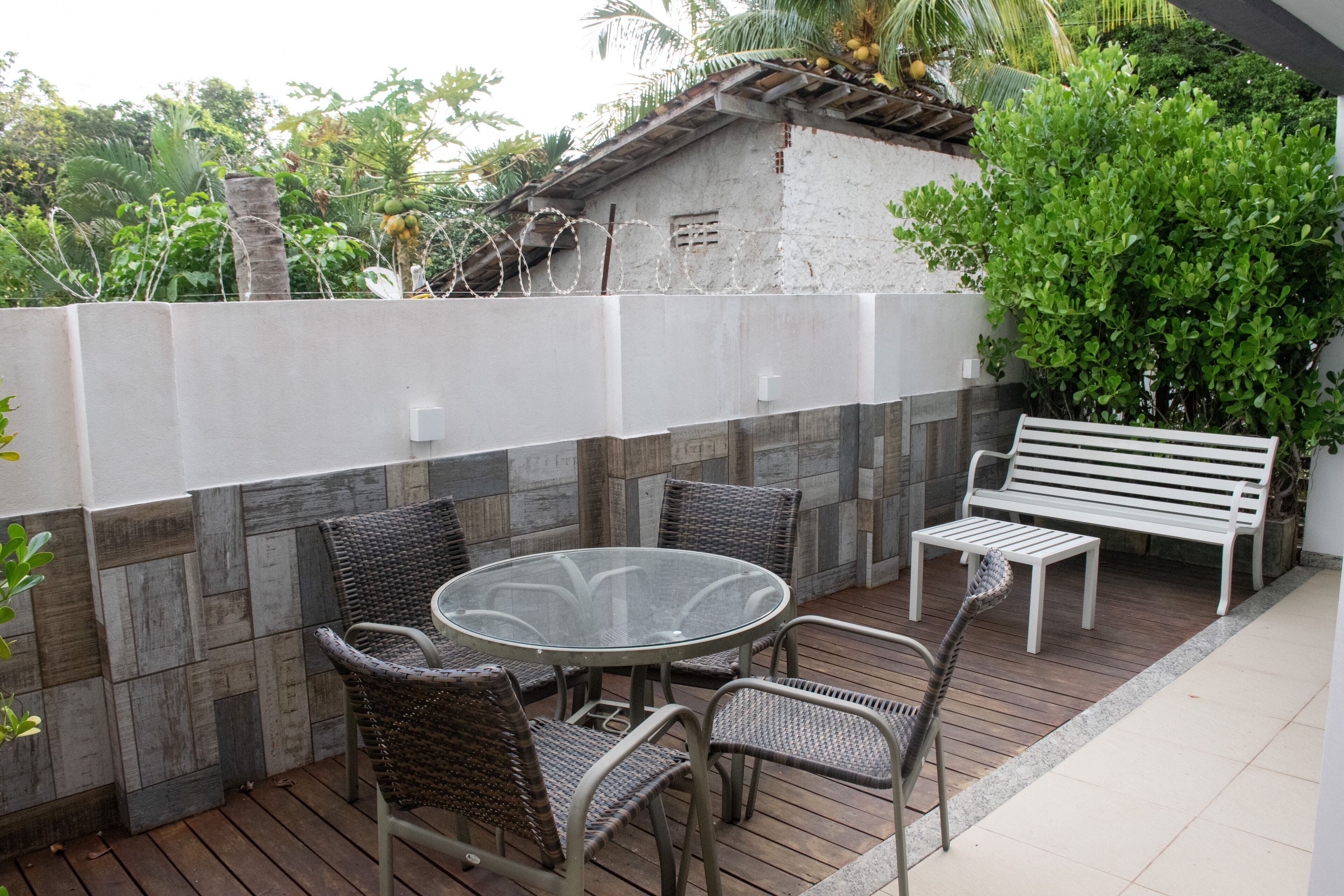 Een terras met een ronde glazen tafel, vier rieten stoelen, een witte bank en een witte muur met prikkeldraad.