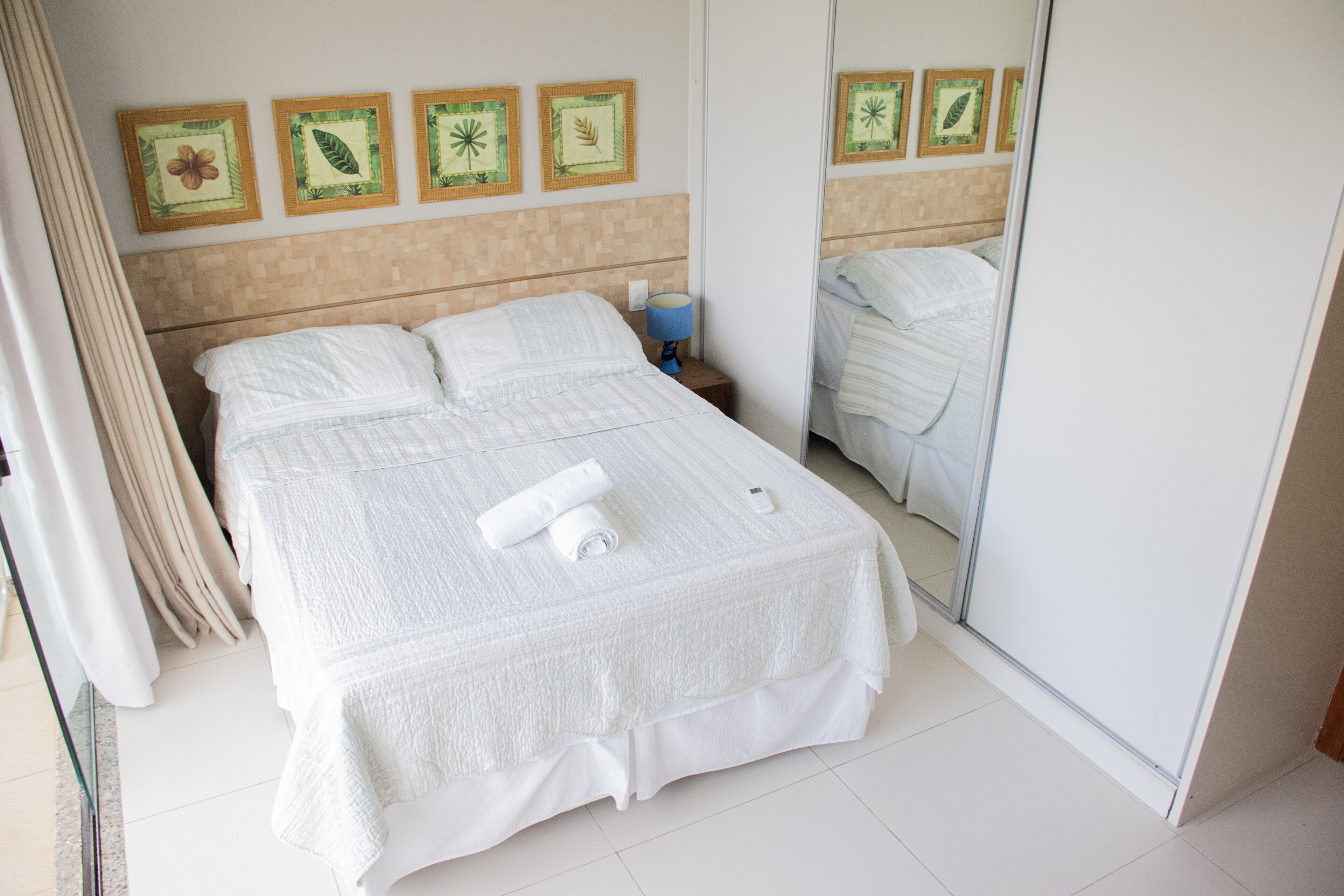 Una cama bien hecha con una colcha blanca y dos almohadas, una lámpara de noche y una pared decorada con impresiones botánicas enmarcadas.