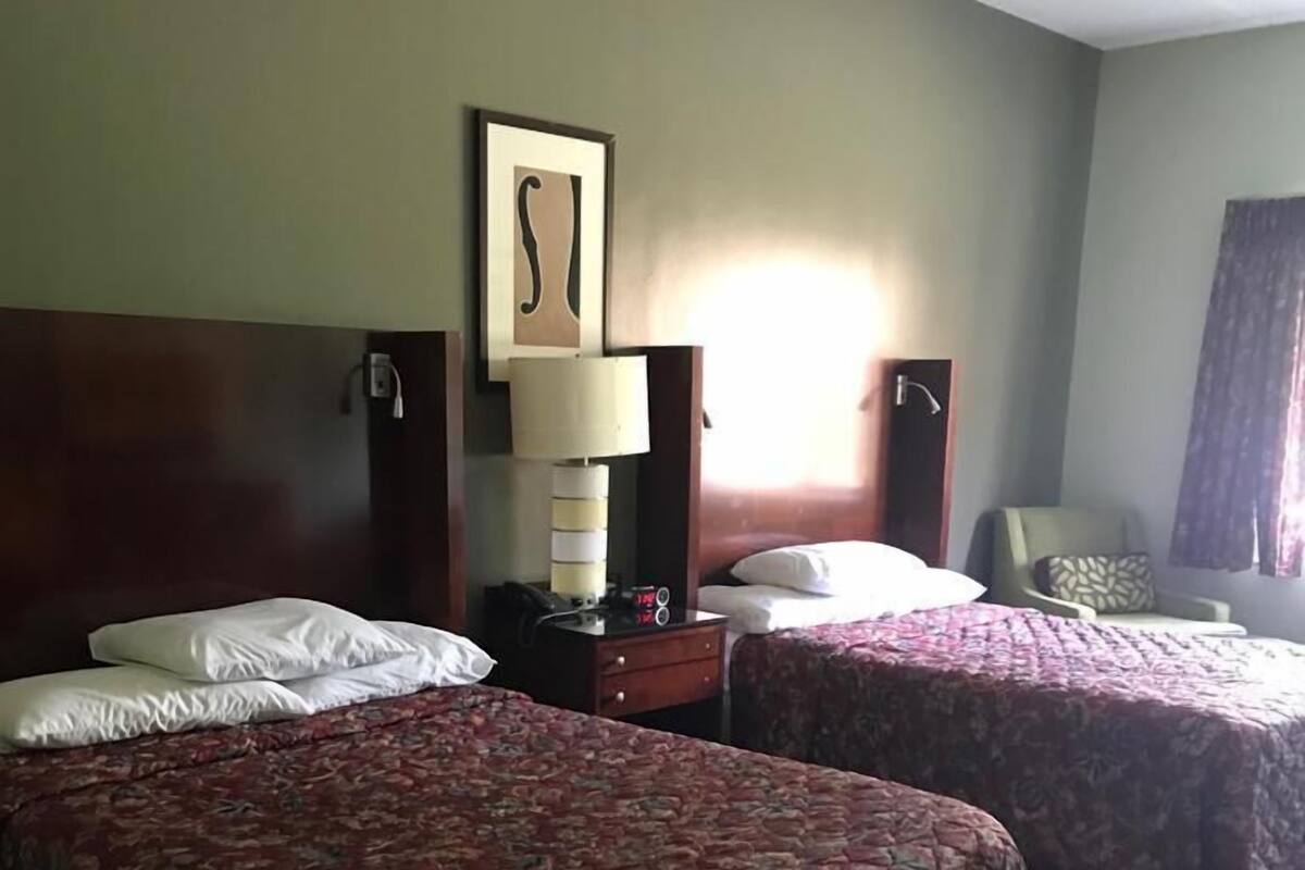 A hotel room with two beds, a bedside table with a lamp, a wall-mounted artwork, and a window with curtains.