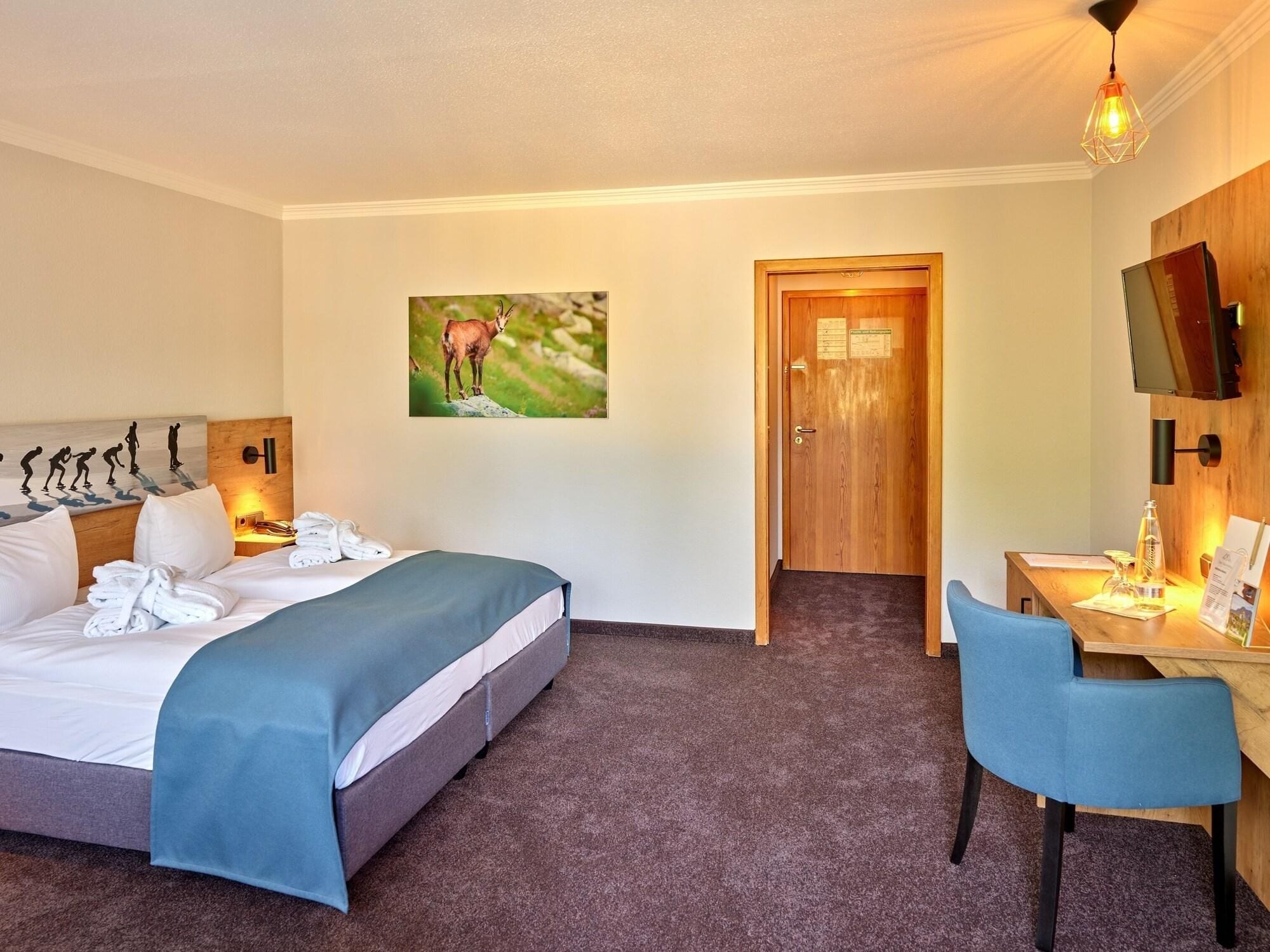 A hotel room with a bed, a desk, a chair, a painting of a deer, and a television on the wall.