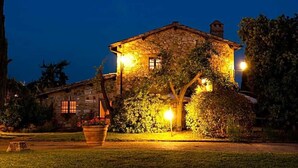 Una casa de piedra con techo inclinado, iluminada por luces cálidas, rodeada de un jardín cuidado y un cielo nocturno despejado.