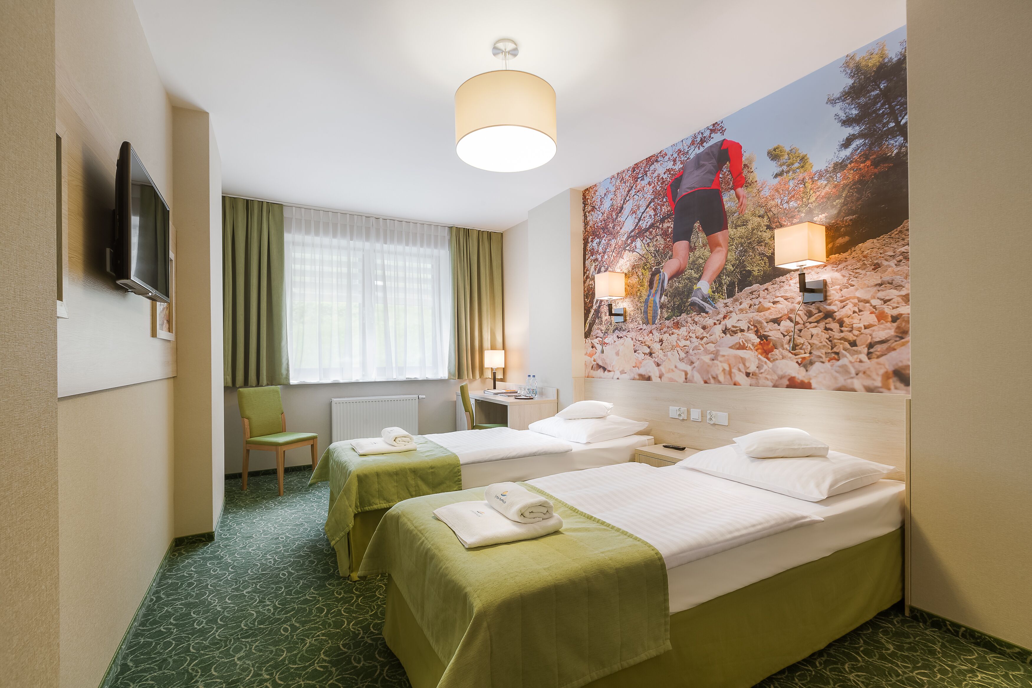 A hotel room with two beds, a television, a desk with a lamp, a chair, and a large painting of a hiker.