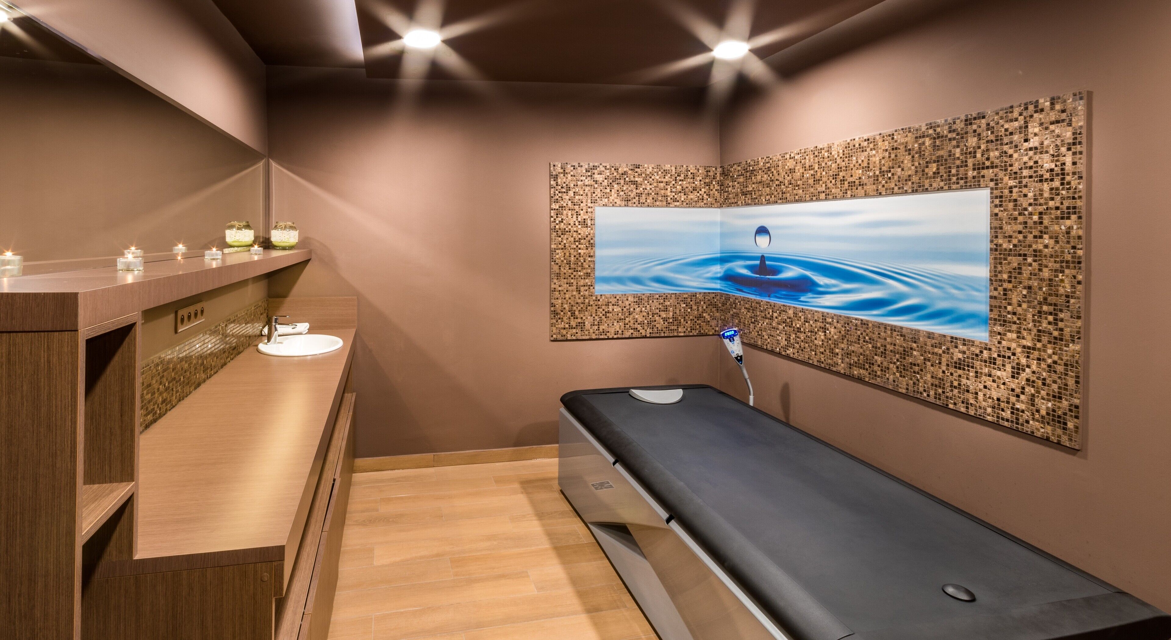 Ein Wellnessraum mit einer Holzbänkl, einem an der Wand befestigten Massagetisch, einem kleinen Waschbecken und einem Wandfernseher, der eine ruhige Wasserszene zeigt.