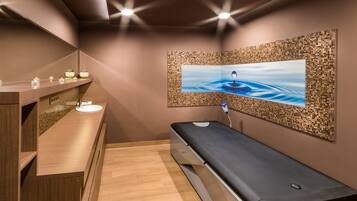 Ein Wellnessraum mit einer Holzbänkl, einem an der Wand befestigten Massagetisch, einem kleinen Waschbecken und einem Wandfernseher, der eine ruhige Wasserszene zeigt.