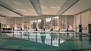 Une grande piscine intérieure avec des parois en verre transparent, de nombreuses fenêtres et des chaises longues disposées le long du bord de la piscine.