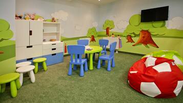 Uma sala de recreação infantil com carpete verde, uma mesa redonda com cadeiras azuis, um armário branco e um puff vermelho e branco.