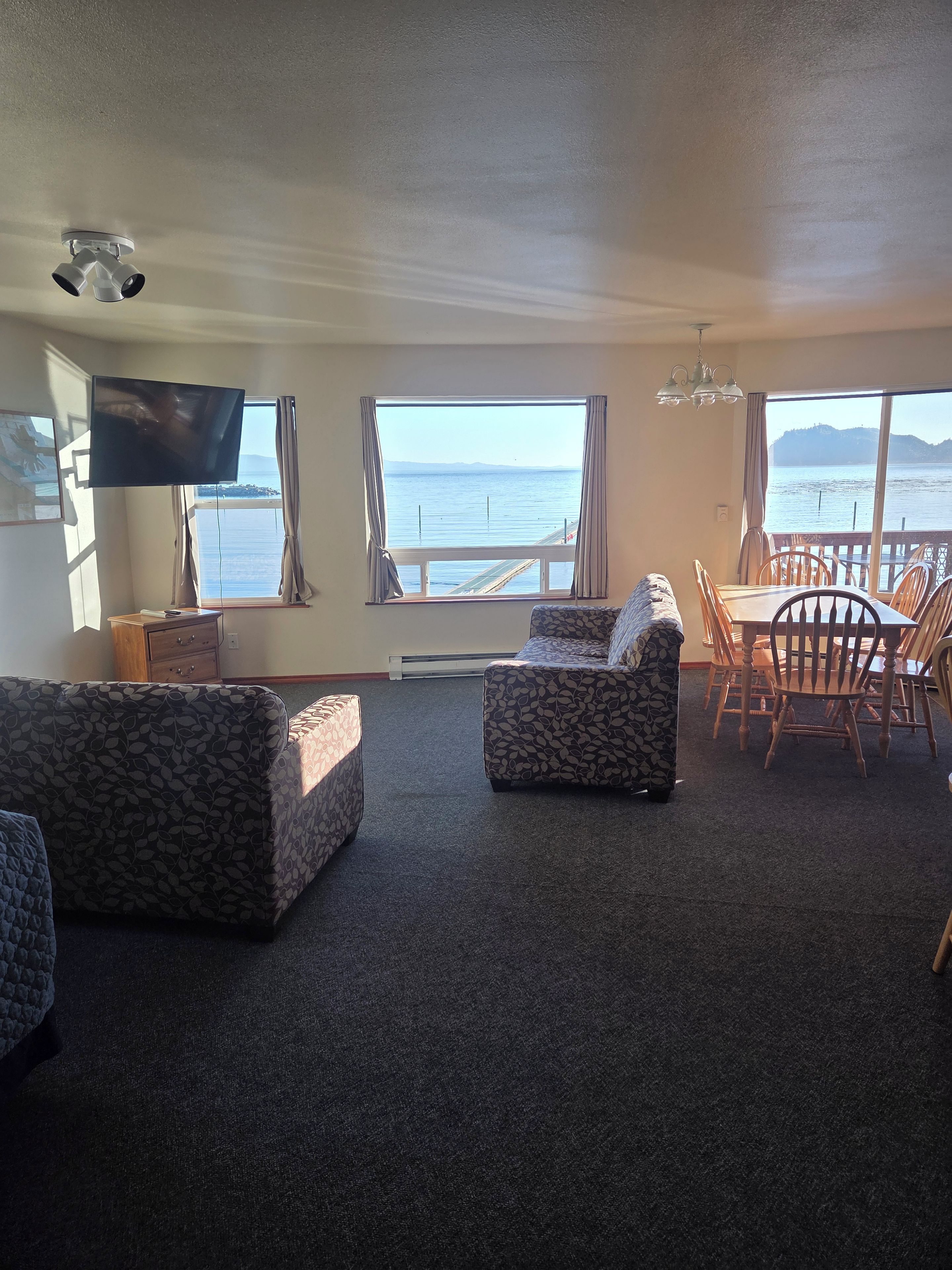 Ein Wohnzimmer mit Fernseher, zwei Sofas, einem Esstisch mit Stühlen und großen Fenstern mit Meerblick.