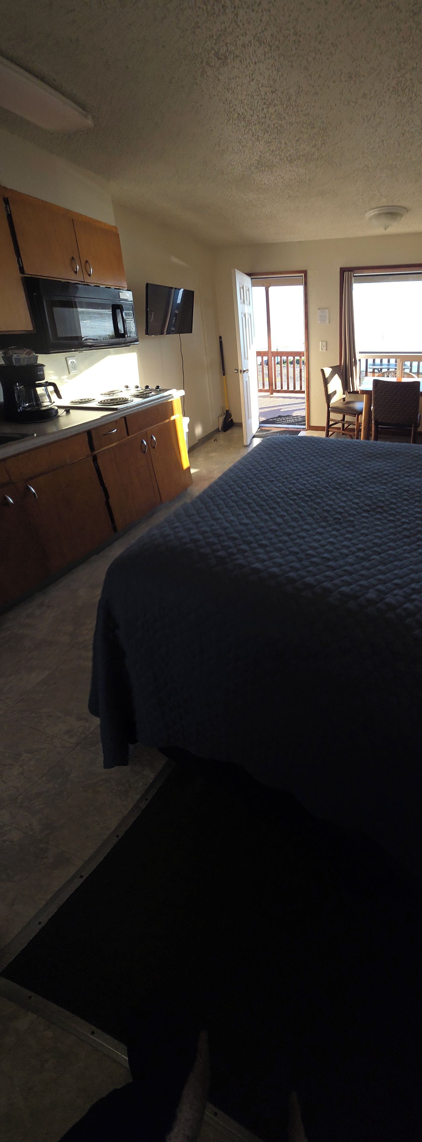 A bedroom with a bed, a desk, a chair, a television, and a kitchenette.