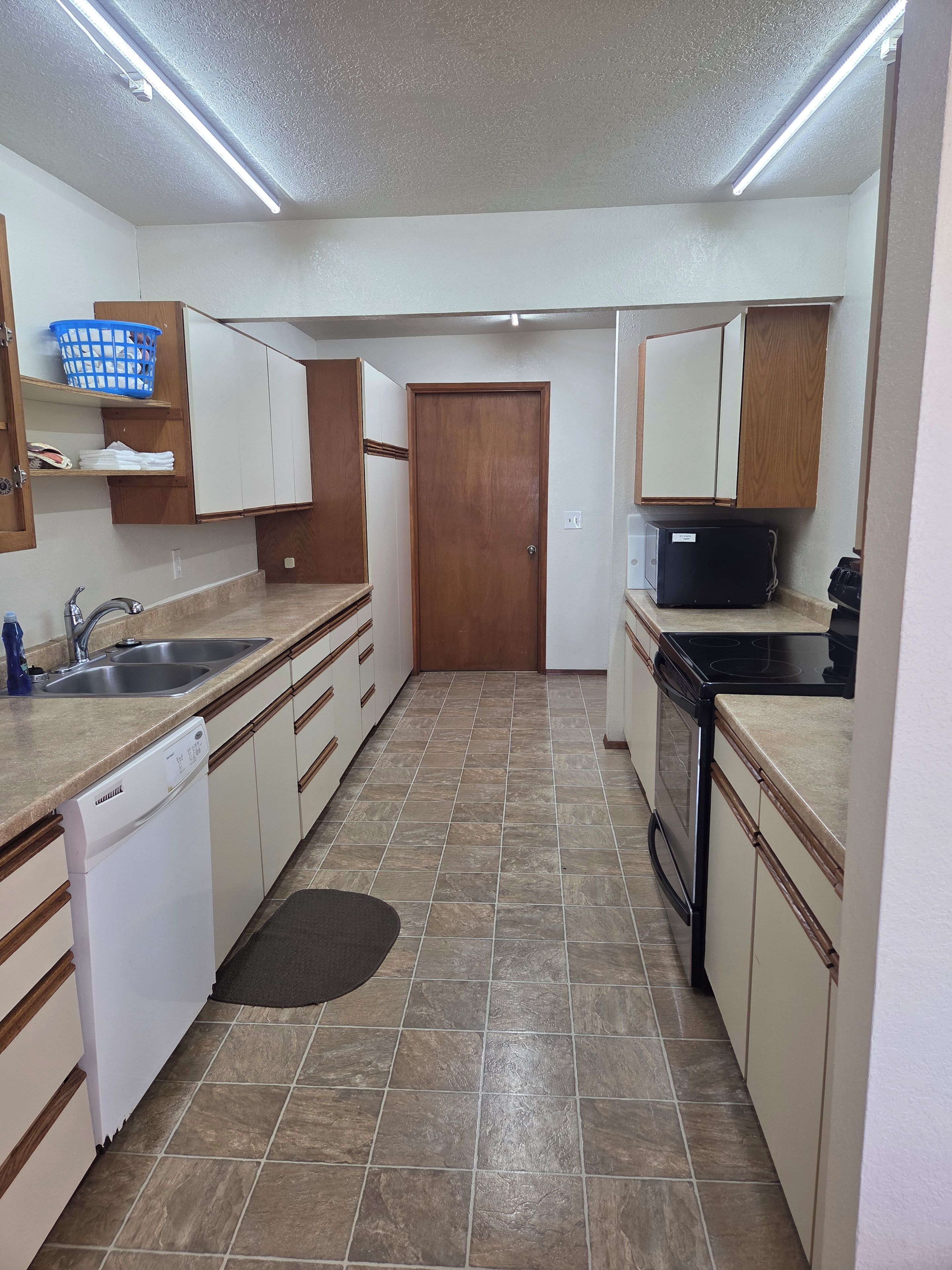A compact kitchen with a dishwasher, stove, and a microwave. There is a wooden door and a blue basket on a shelf.