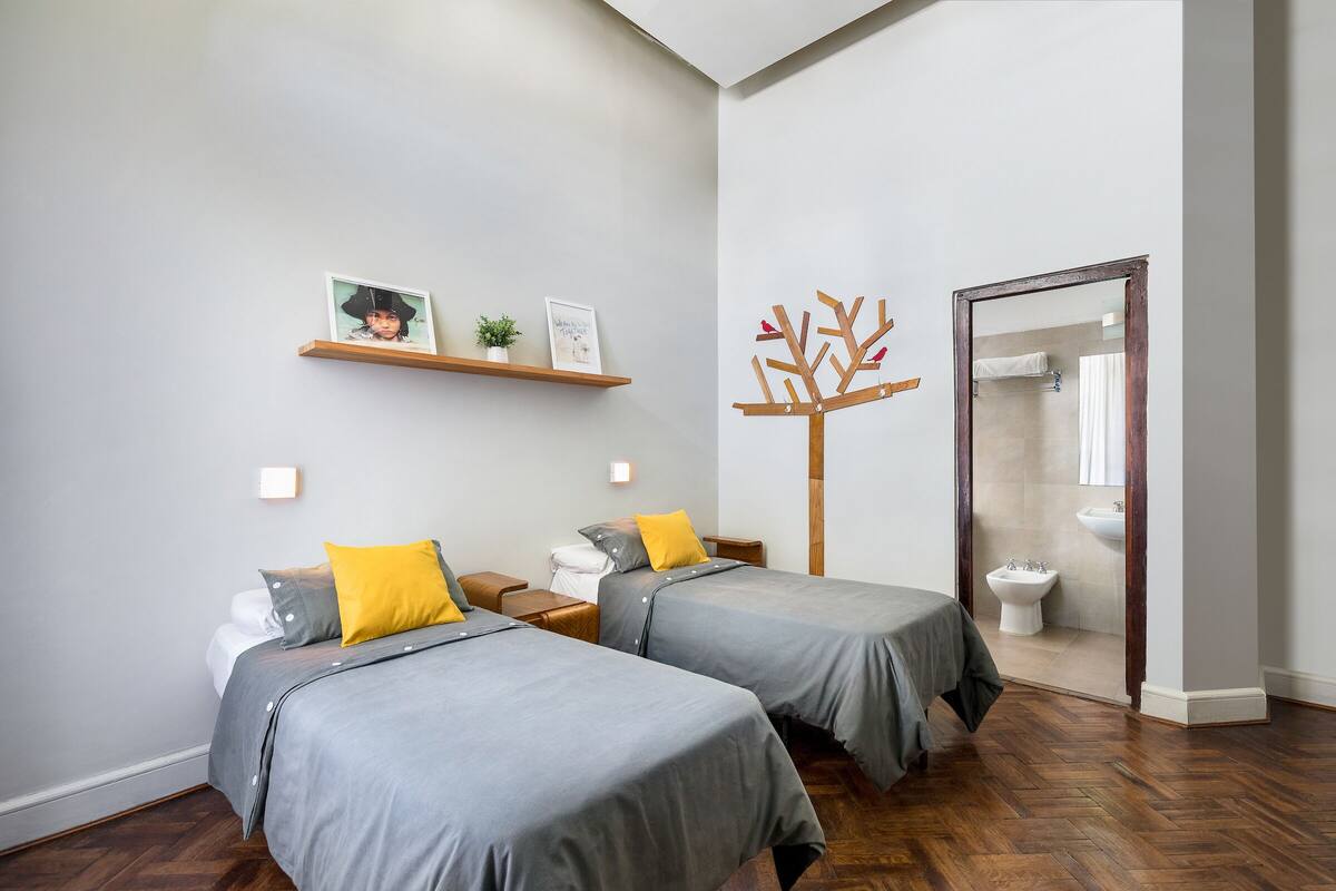 Un dormitorio moderno con dos camas, suelo de madera, un baño visible a través de un vidrio y un estante pequeño con fotografías.