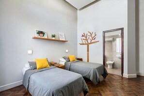 Un dormitorio moderno con dos camas, suelo de madera, un baño visible a través de un vidrio y un estante pequeño con fotografías.