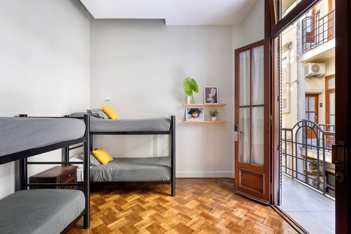 Una habitación compacta con literas, suelo de madera en patrón espiga, una mesita de noche pequeña, un estante con una planta e imagen, y una puerta que da a un balcón con reja de metal.