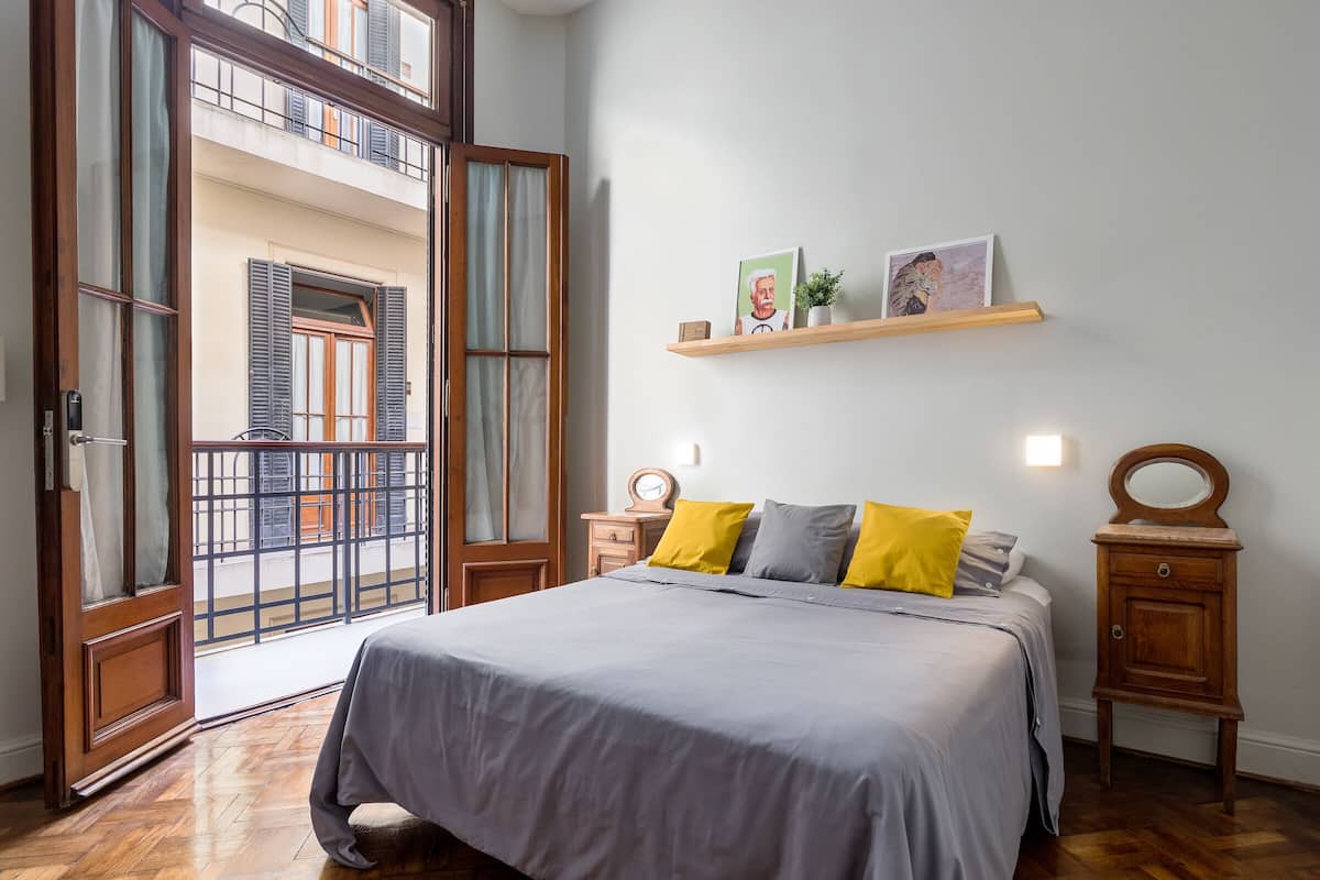 Un dormitorio con cama, mesitas de noche de madera, un estante con fotografías y un balcón con contraventanas.