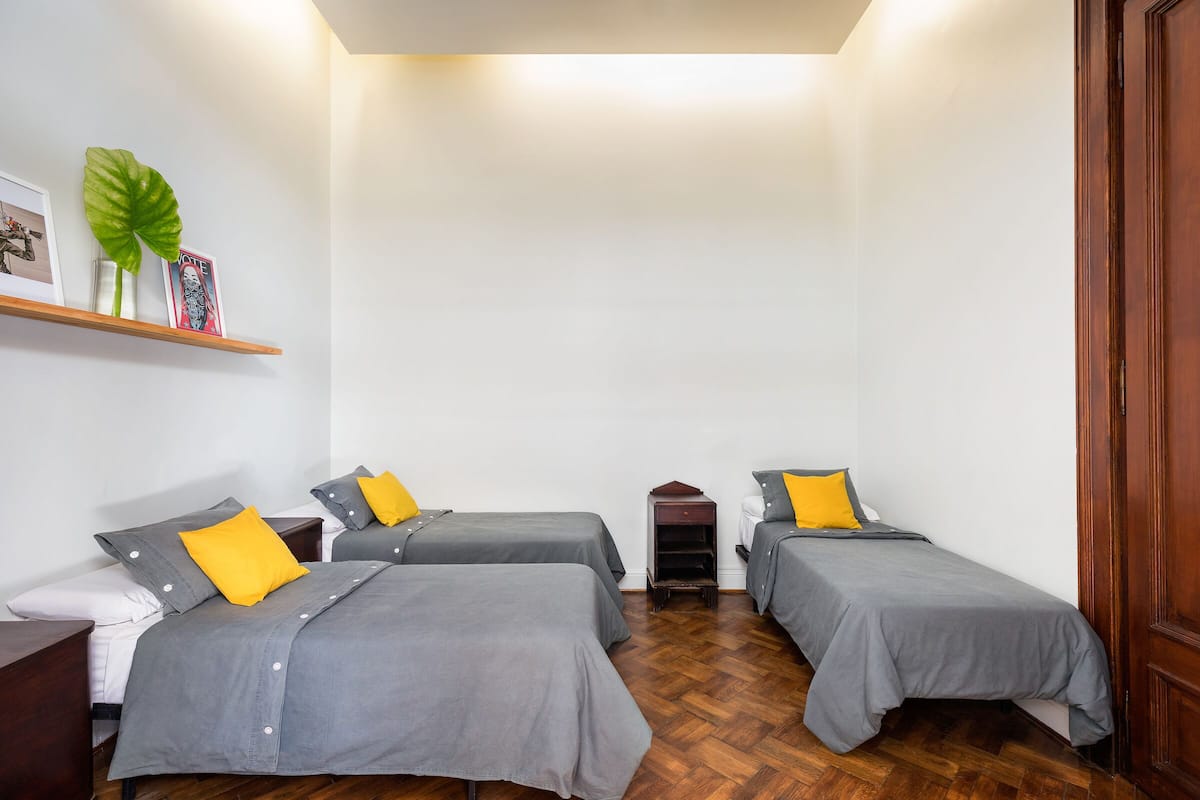 Una habitación pequeña y ordenada con tres camas, cada una con cubrecama gris y almohada amarilla. Hay un estante de madera con una planta y un cuadro enmarcado. El suelo es de madera y la puerta también.