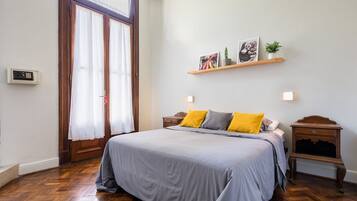 Un dormitorio con una cama grande, suelo de madera, una puerta con cortinas, una mesita de noche y una estantería con fotos y una planta.