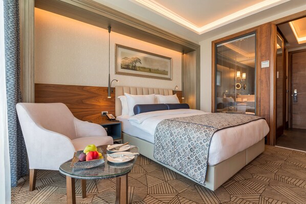 A hotel room with a bed, a chair, a small table with fruit, and a painting on the wall.
