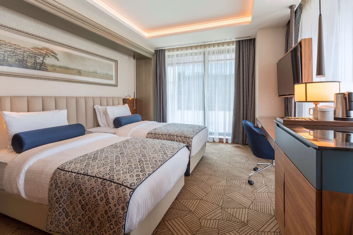 Une chambre d’hôtel avec deux lits, un bureau, une chaise, une télévision et une grande fenêtre avec des rideaux.
