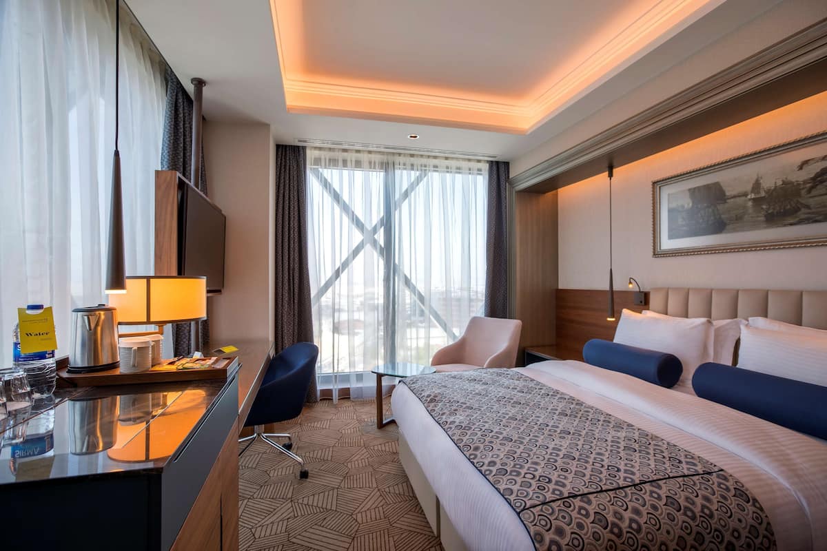 Une chambre d’hôtel comprenant un lit, un bureau avec une machine à café, une chaise, un téléviseur et une grande fenêtre avec des rideaux.