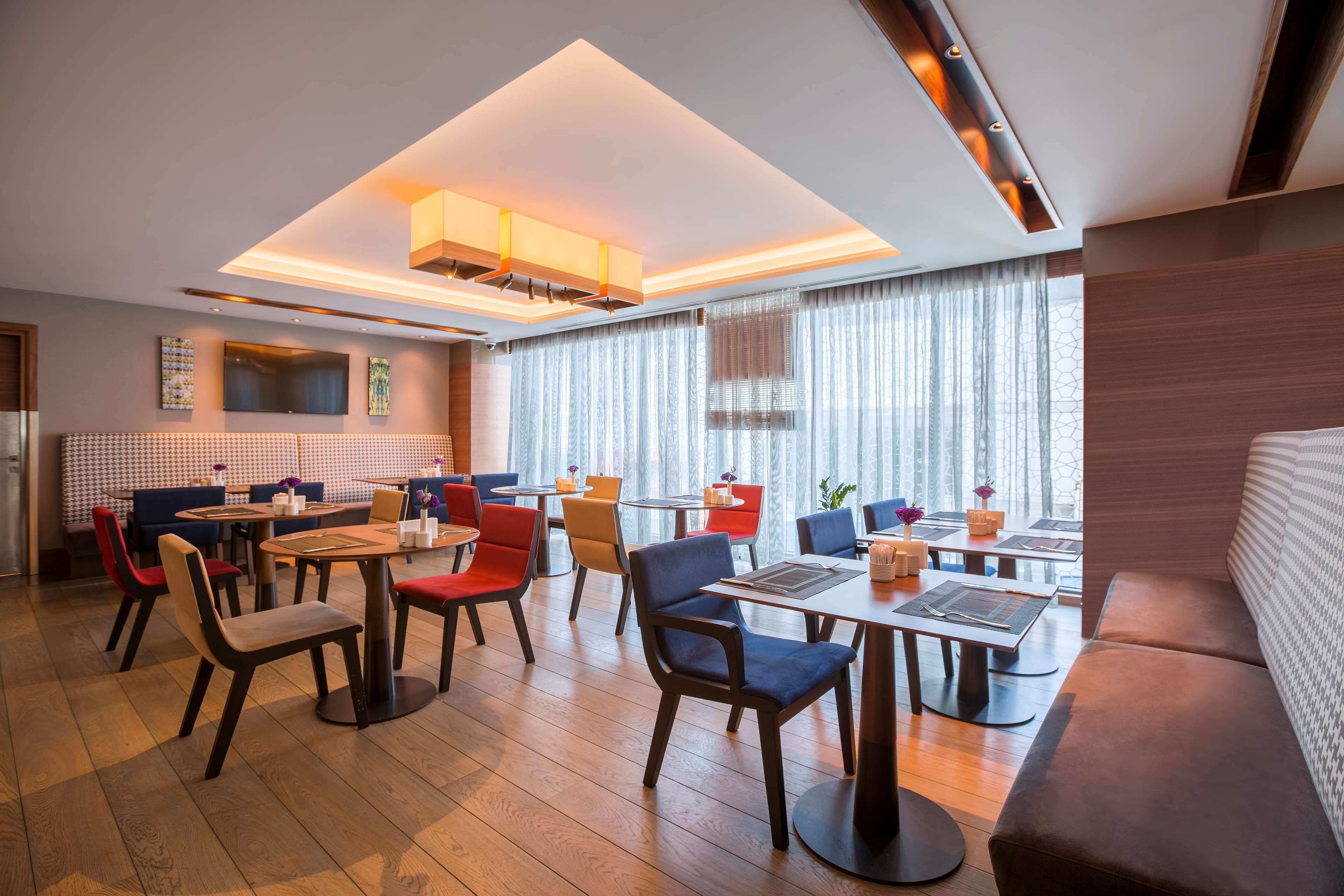 A modern dining area with multiple tables and chairs, a large window with curtains, and a ceiling with recessed lighting.