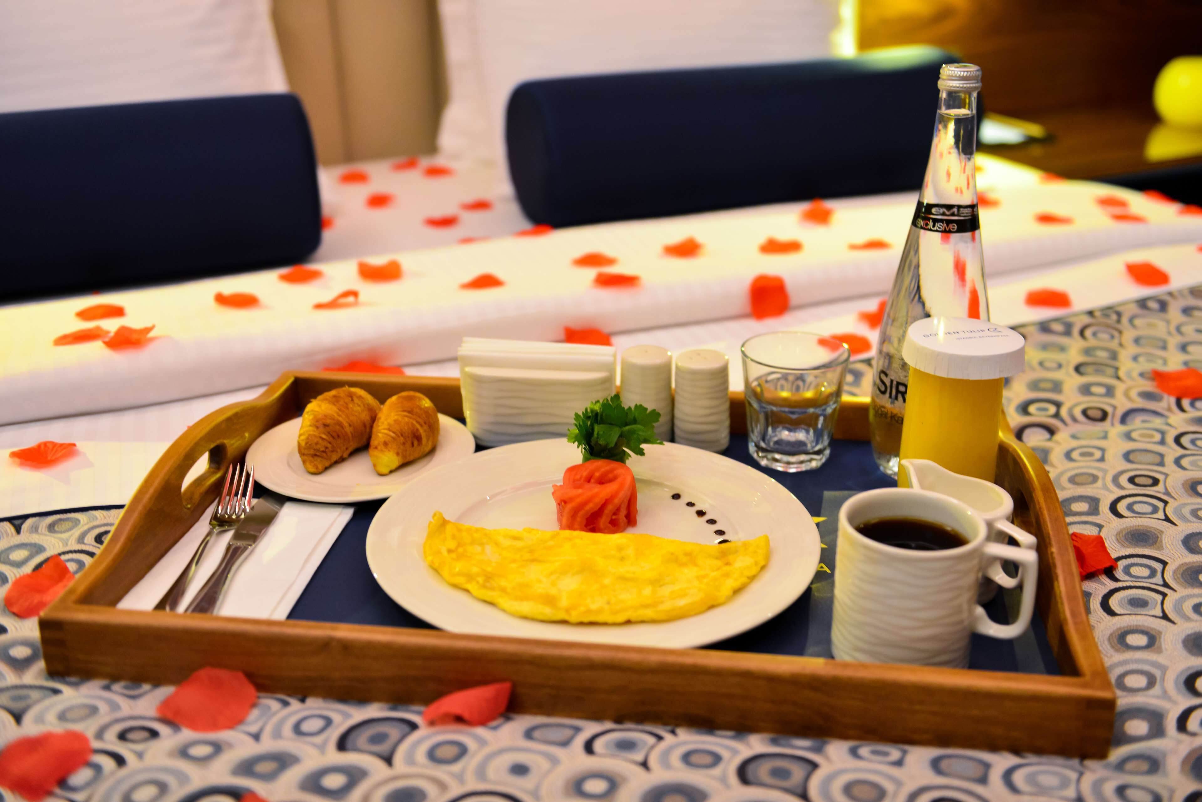 A breakfast tray with croissants, an omelet, a small salad, a bottle of champagne, a cup of coffee, and a glass of water on a bed with rose petals.