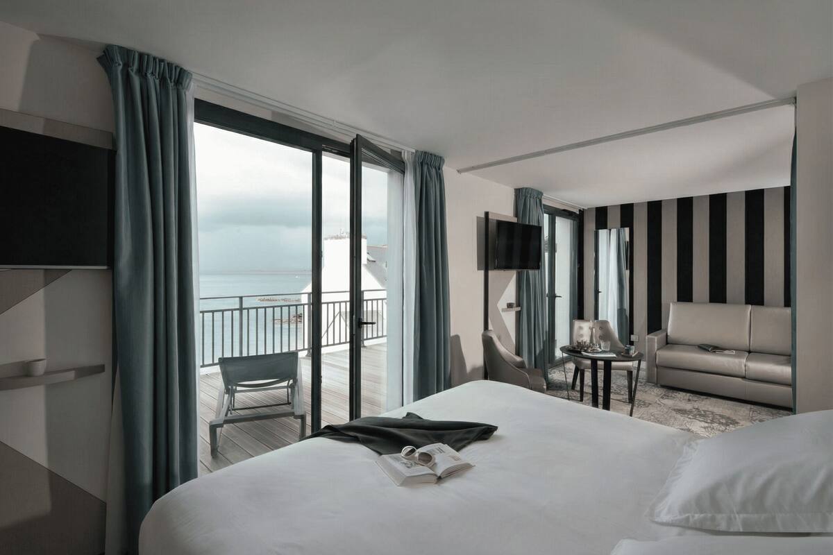 Ein Hotelzimmer mit Bett, Sofa, Fernseher, kleinem Tisch und einem Balkon mit Meerblick.