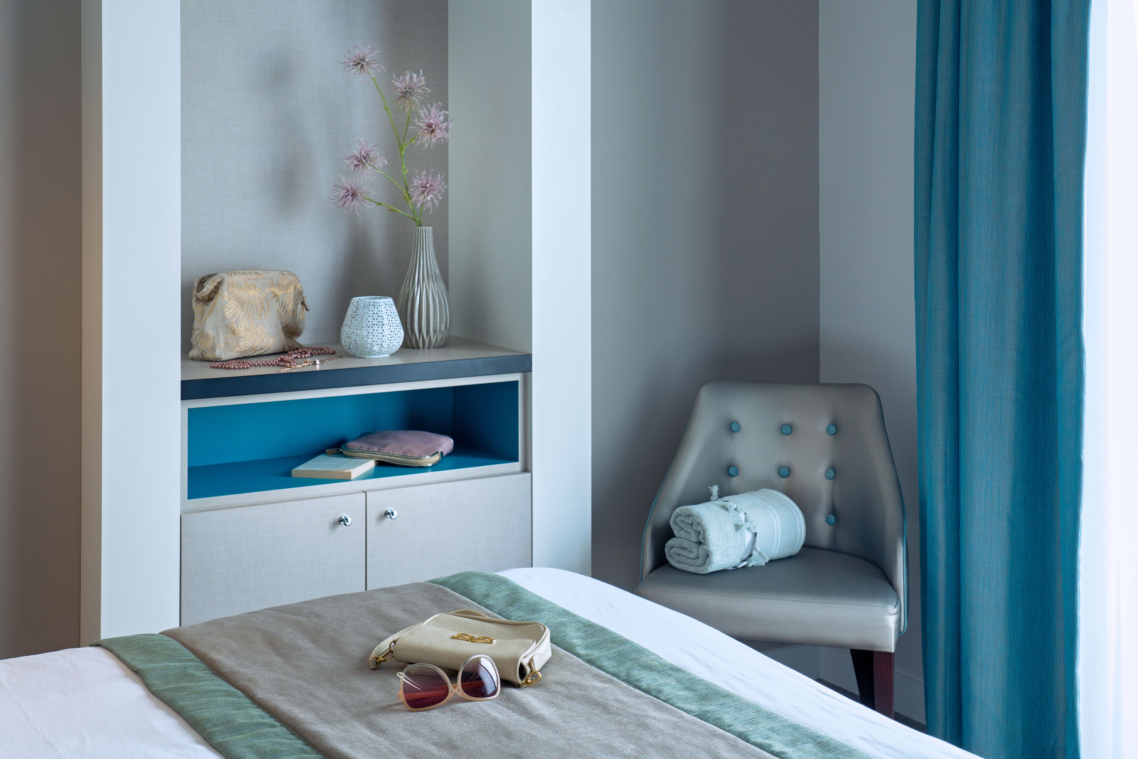 Habitación de hotel impecable con cama, una silla, estante con adornos y ventana con cortinas.