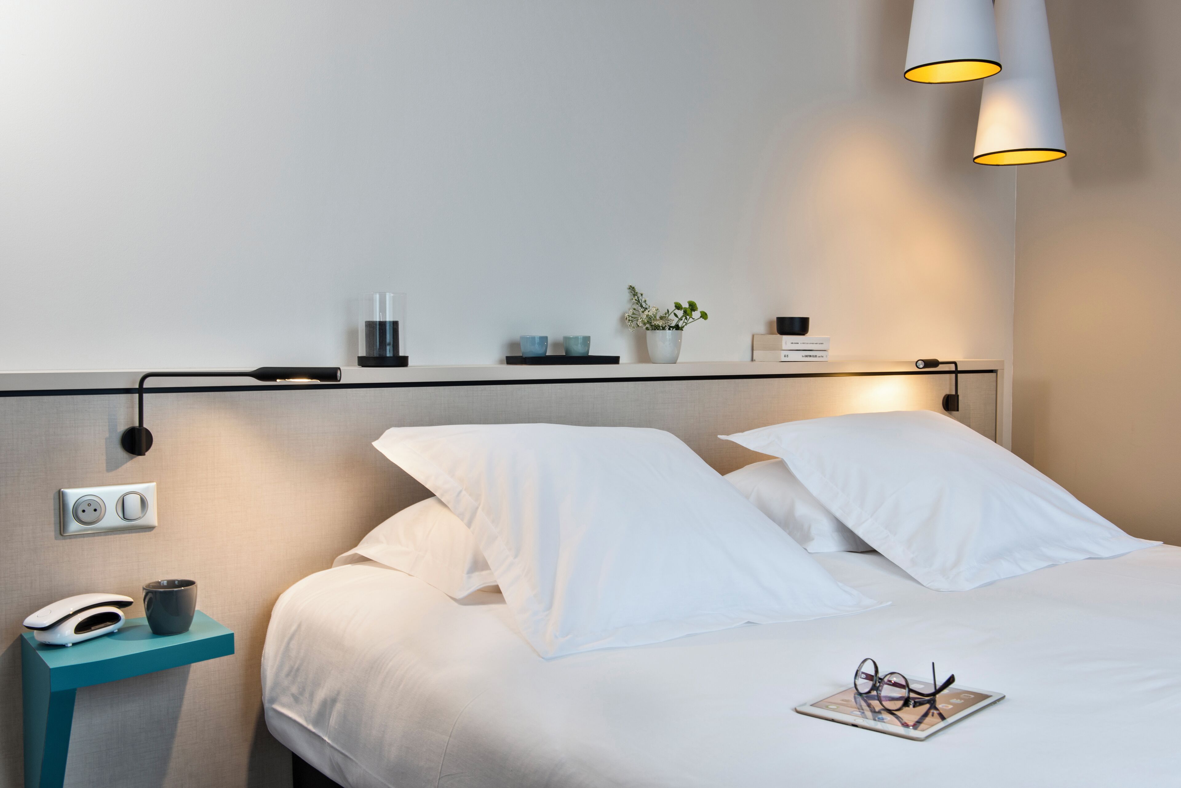 A neatly made bed with white linens, a bedside table with a lamp, a phone, and a cup.