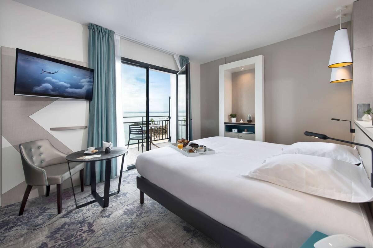 Ein modernes Hotelzimmer mit einem großen Bett, einem Schreibtisch mit Stuhl, einem an der Wand befestigten Fernseher und einem Balkon mit Blick auf den Himmel.