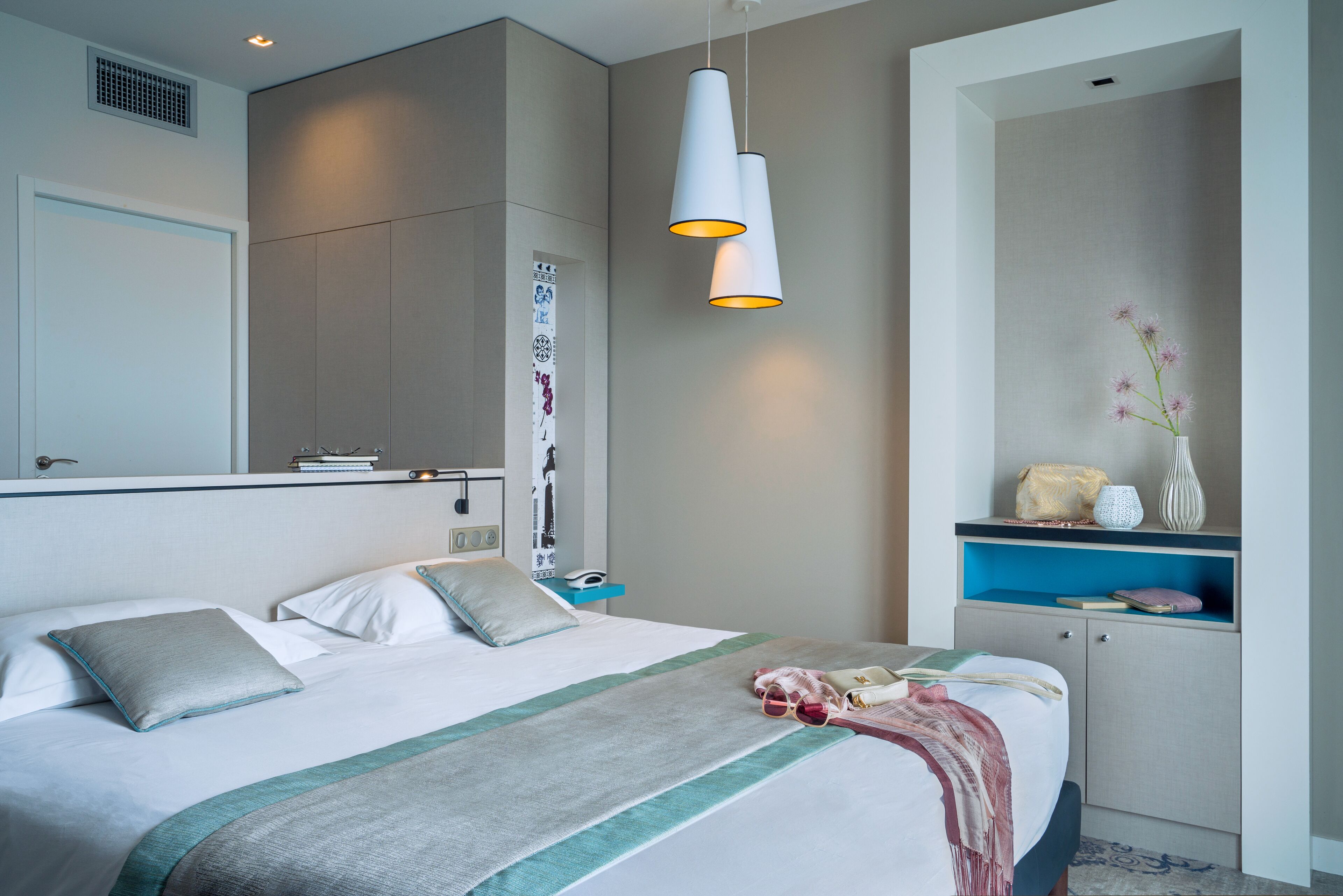 A modern hotel room with a large bed, bedside tables, a wardrobe, and a hanging light fixture.