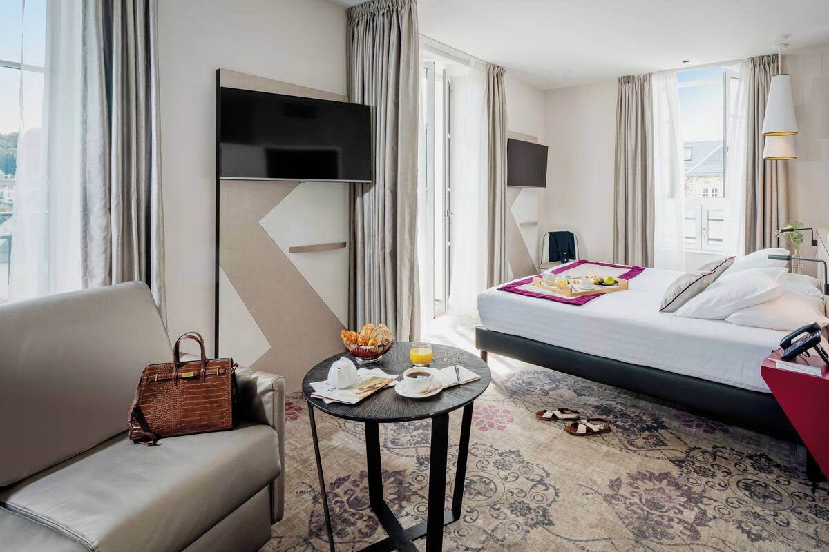 Ein Hotelzimmer mit Bett, Sofa, kleinem Tisch mit Frühstücksangeboten, Fernseher und einem Fenster mit Vorhängen.
