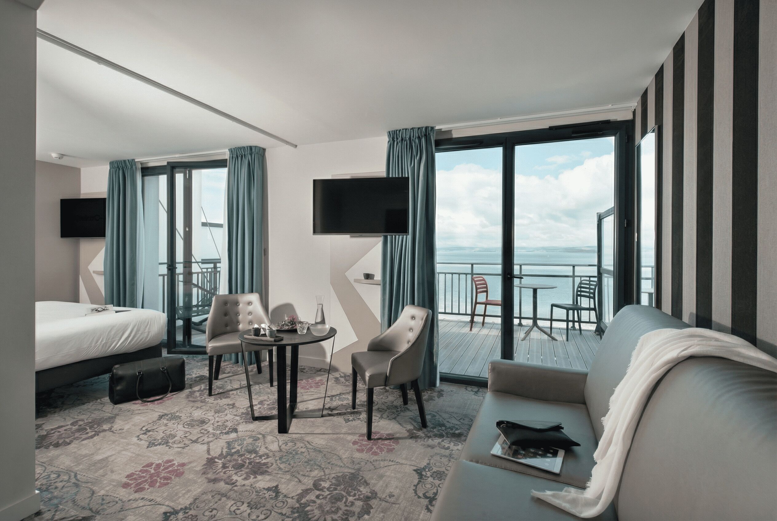 A hotel room with a bed, a sofa, a dining table with chairs, a TV, and a balcony with a view.