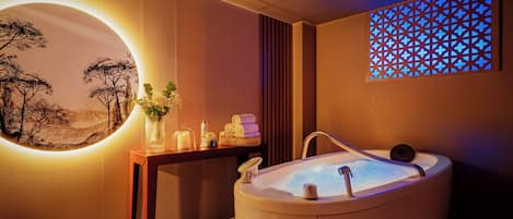 Un cuarto de spa con una bañera grande, un espejo redondo con una pintura de paisaje, una pequeña mesa con velas y flores, y un panel decorativo en la pared.