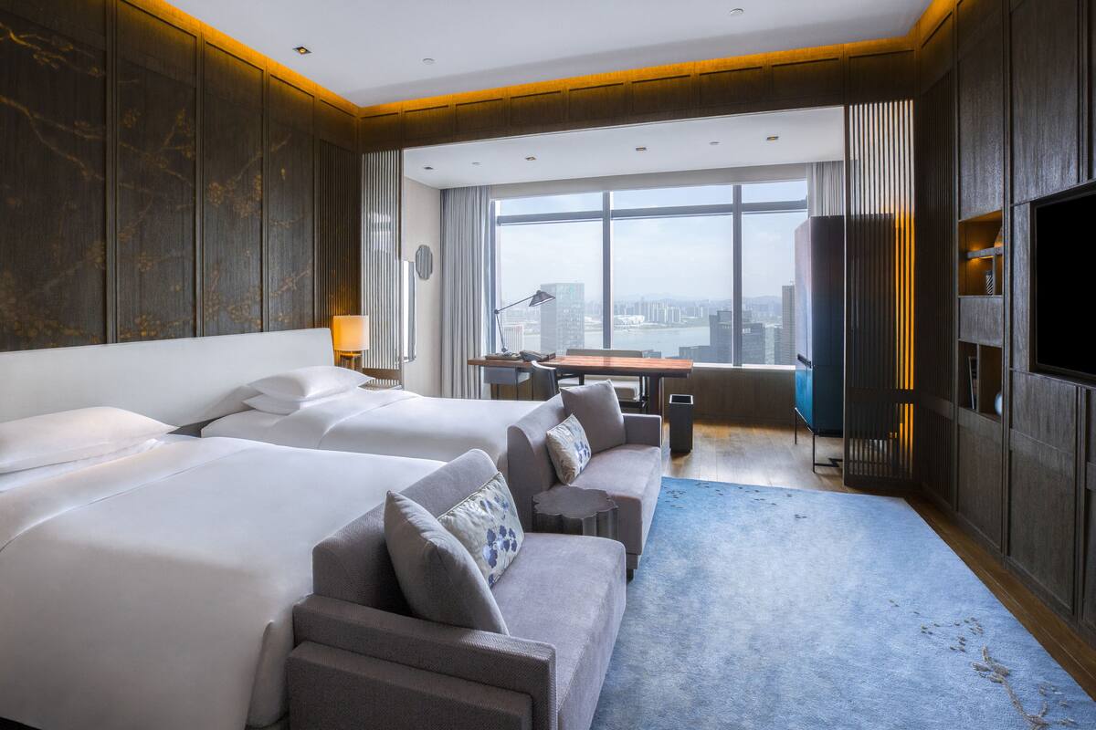 Habitació d'hotel amb dos llits, un sofà, escriptori i una finestra gran amb vista a la ciutat.