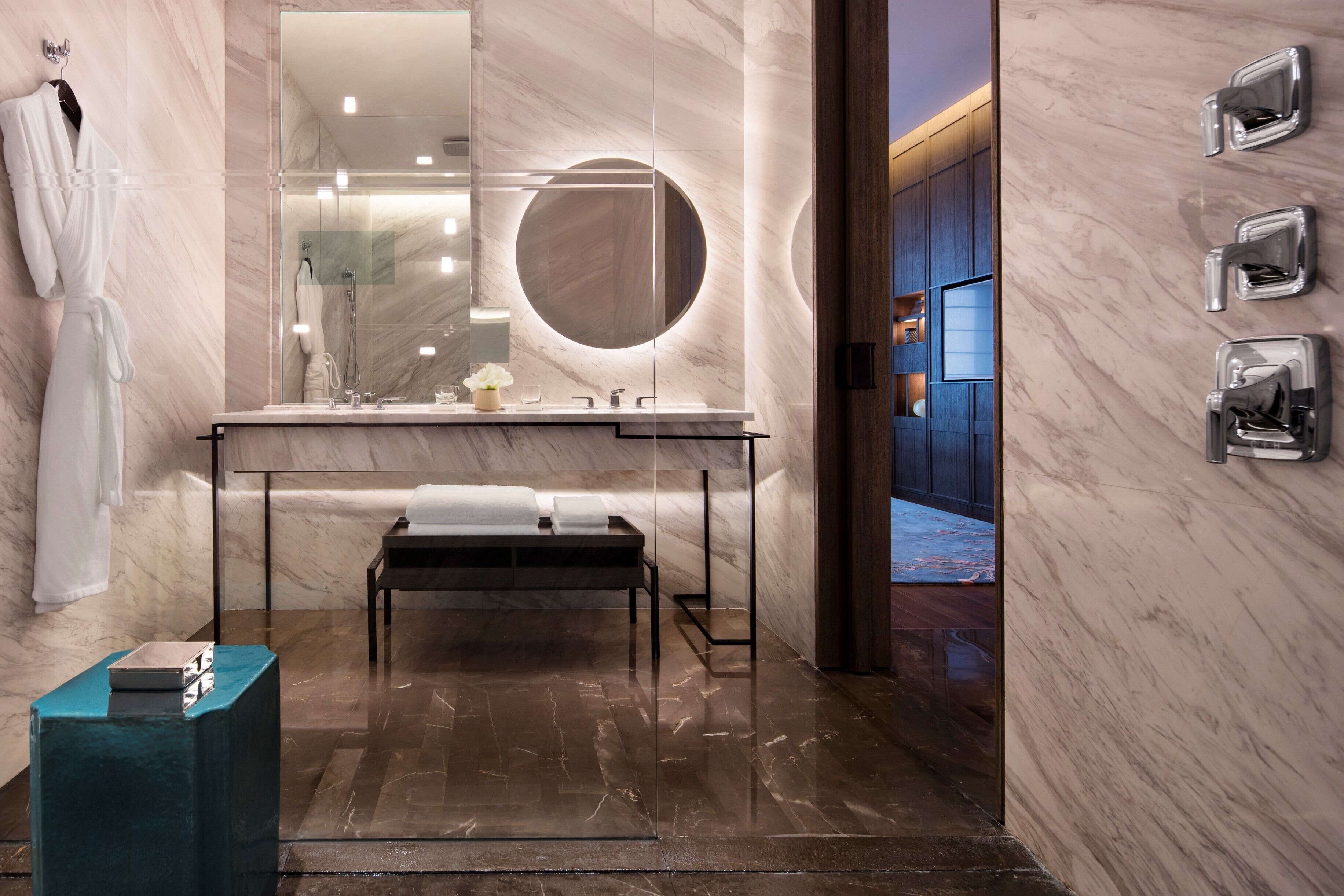 A modern bathroom with a marble vanity, a round mirror, a bench, and a robe hanging on the wall.
