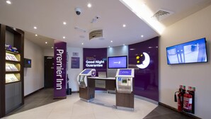 A hotel lobby with a reception desk, a digital check-in kiosk, a television screen, a fire extinguisher, and a sign indicating a 'Good Night Guarantee'. - Premier Inn London Brixton (London)