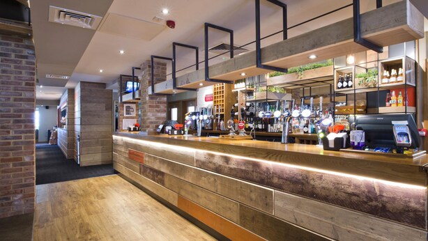 A bar area with a counter, bar stools, and a variety of bottles and glasses. - Premier Inn Farnham (Farnham)