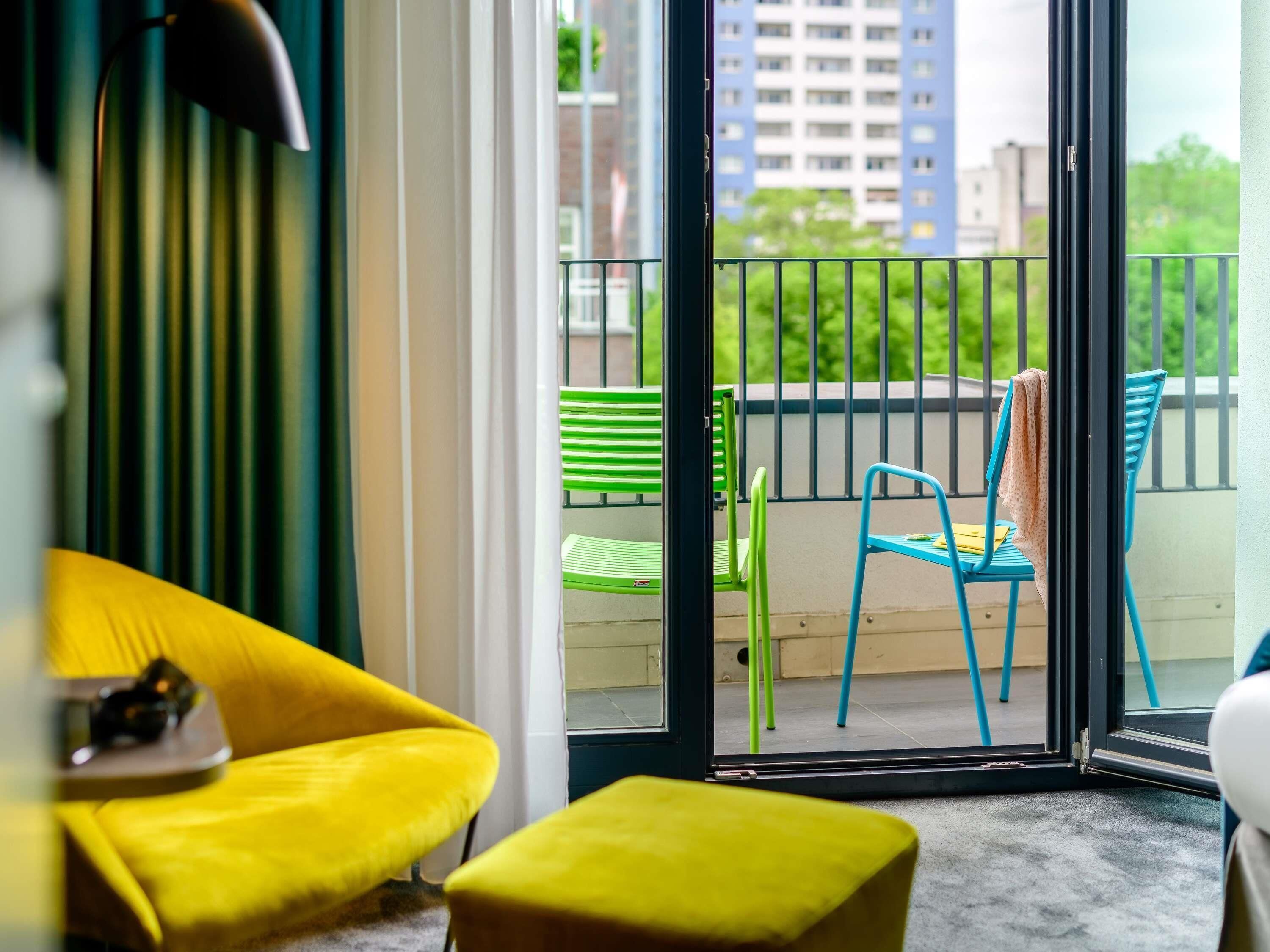 A room with a yellow sofa, a green chair, and a balcony with a view of buildings and greenery.