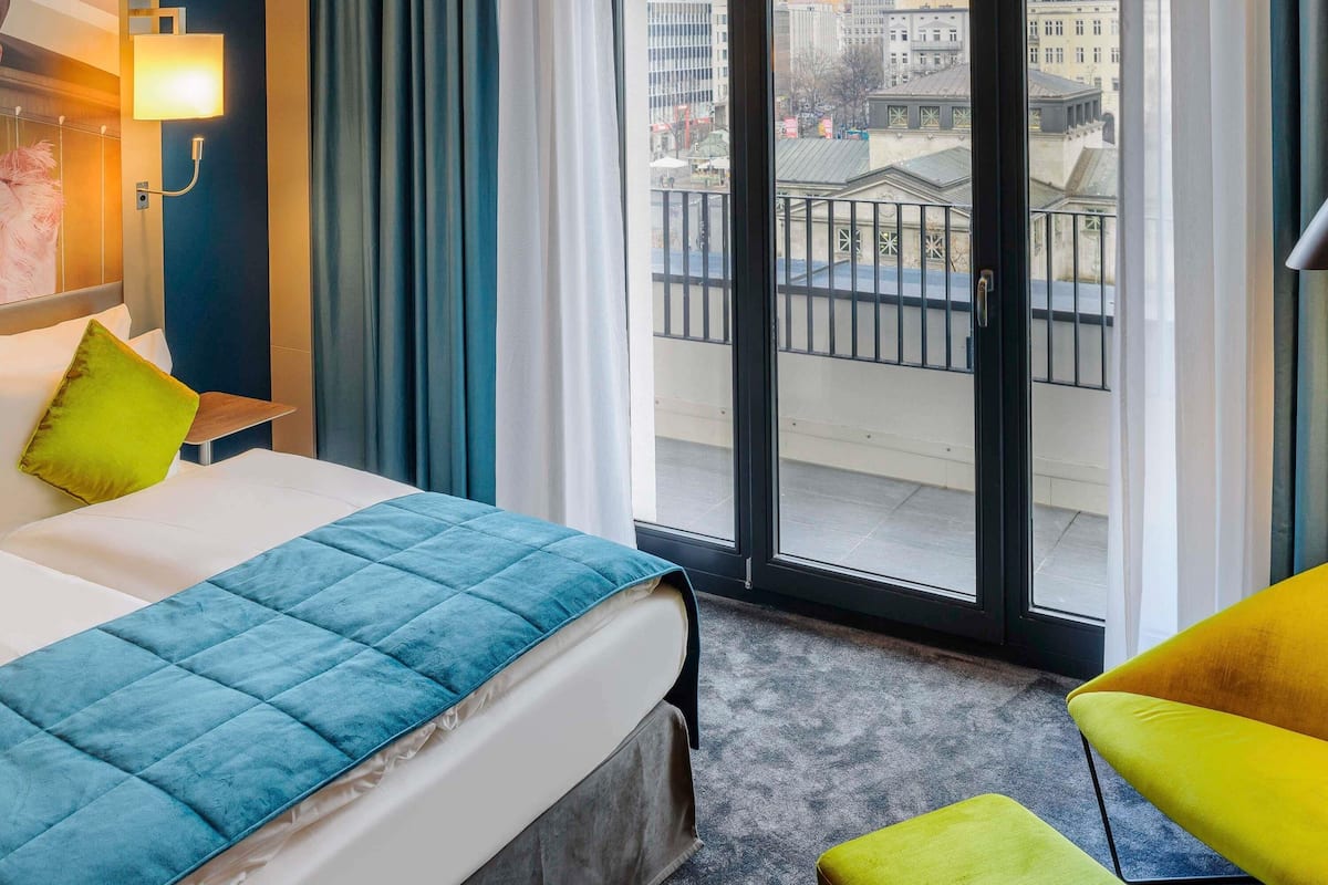 Ein Hotelzimmer mit Bett, Sessel, Fenster und Blick auf Gebäude sowie einer Tür.