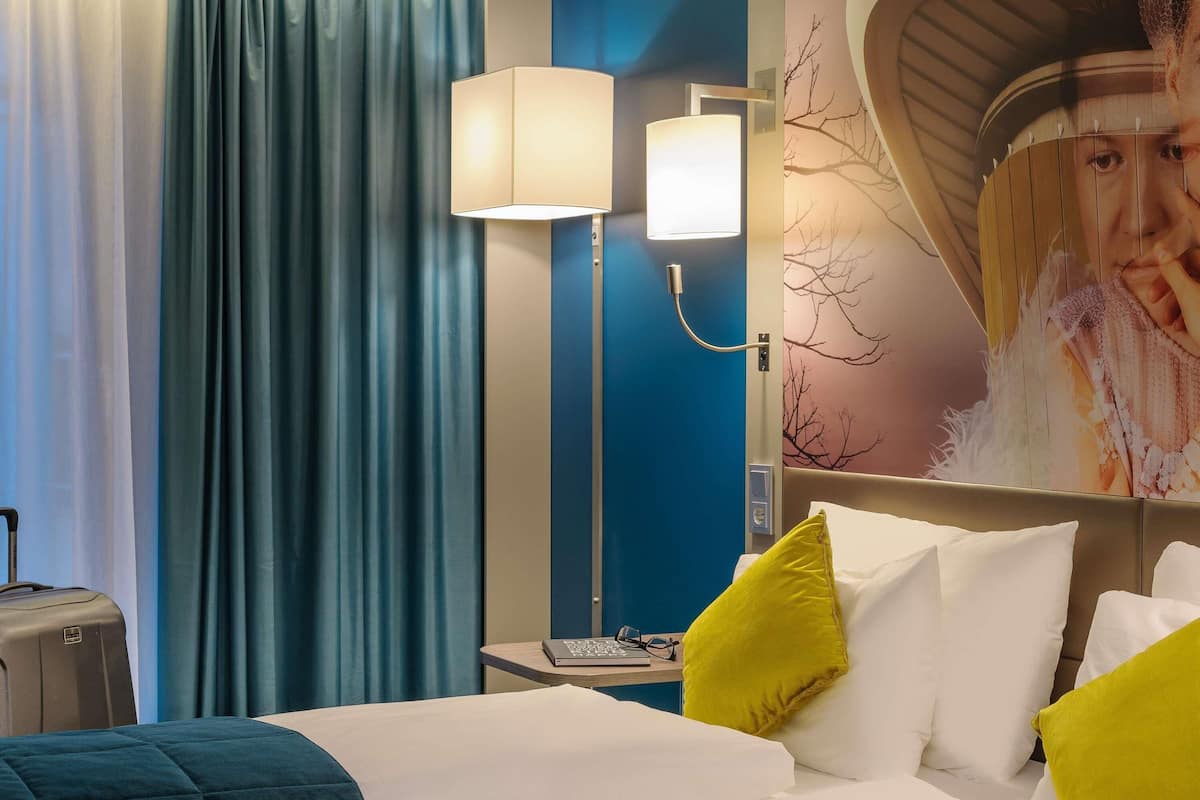 Ein Hotelzimmer mit Bett, Schreibtisch, Laptop, Lampe und einer Wand mit Baum-Wandmalerei.