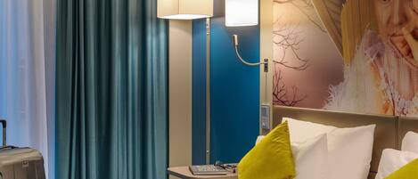 A hotel room with a bed, a desk with a laptop, a lamp, and a wall with a tree mural.