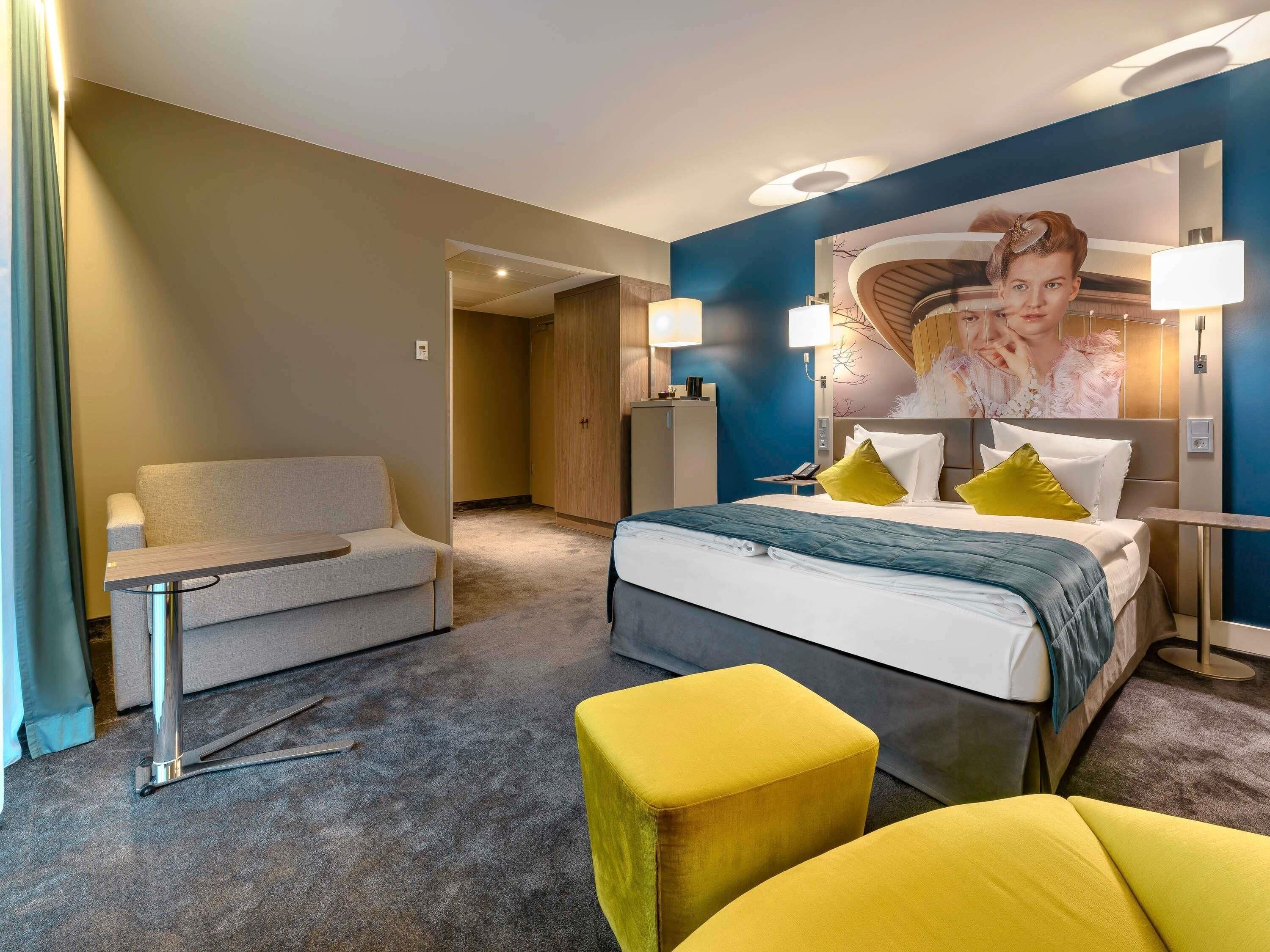 Une chambre d’hôtel avec un lit, un canapé, une petite table et un pouf jaune.