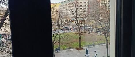 Blick aus einem Fenster auf eine Stadtstraße mit Gebäuden, einem parkenden Auto und Fußgängern.