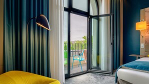 Une chambre d’hôtel avec un lit, un fauteuil jaune, une chaise bleue, une lampe et une porte-fenêtre coulissante donnant sur un espace extérieur avec une chaise et une clôture.